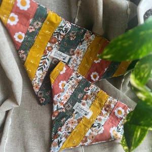 Peut inclure: Deux bandanas pour chiens avec un motif patchwork. Les bandanas présentent des rayures verticales dans les tons de jaune, corail, vert et des motifs floraux. Un bandana est plus grand que l'autre. Les bandanas ont le texte "Patchwork Pups Co."