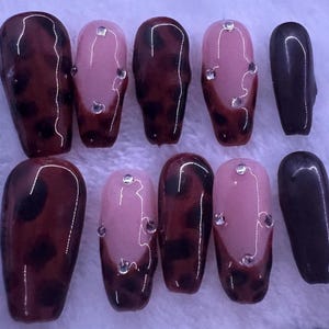 May include: A set of ten artificial nails in various designs. Some nails feature a dark brown leopard print, others are light pink with rhinestones, and a few are solid dark brown.