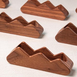 May include: Several wooden mountain-shaped pen holders are arranged on a white surface. The pen holders are made of a dark brown wood and have a carved mountain range design. The pen holders are designed to hold pens or pencils.