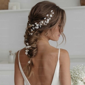 May include: A delicate floral hair vine with white blossoms and pearl accents, adorning a braided hairstyle. The hair accessory is designed for bridal or special occasion wear, adding a touch of elegance and romance to the look.