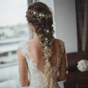 May include: A bride's hairstyle featuring a braided updo adorned with delicate white floral hair accessories. The back of the bride's ivory lace wedding dress is visible, with intricate floral lace detailing. The hairstyle includes a floral headpiece and hairpins.