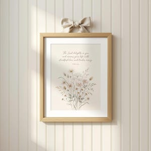 Può includere: Una stampa incorniciata con un design floreale ad acquerello e il testo "The Lord delights in you and crowns your life with steadfast love and tender mercy." La cornice è in legno chiaro, con un fiocco beige in alto.