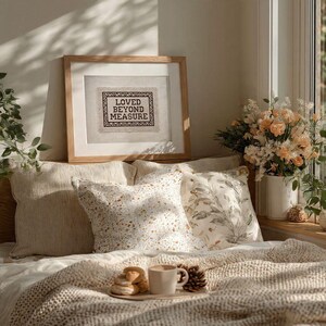 May include: A cozy bedroom scene with a framed artwork that reads "LOVED BEYOND MEASURE". The artwork is above pillows and a bed with a knitted blanket. A tray with a mug, cookies, and a pinecone sits on the bed.