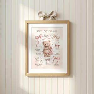 Puede incluir: Una impresión enmarcada con el texto "GOD SAYS I AM" y un oso de peluche sosteniendo zapatillas de ballet. La impresión presenta lazos rosas y beige y las palabras "Fuerte", "Precioso", "Hermoso", "Nunca solo", "Amado", "Valiente", "Elegido" y "Valioso".