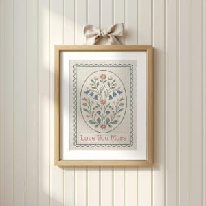 May include: A framed cross-stitch artwork featuring an oval floral design in pastel colors. The design includes bluebell-like flowers and other floral elements. The text "Love You More" is stitched at the bottom. A beige bow is attached to the top of the frame.