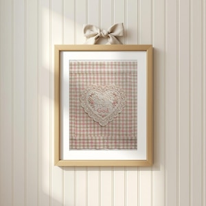 May include: A framed artwork featuring a heart-shaped lace applique with the text "A gift from God" embroidered on a pink and tan gingham background. The artwork is in a light wood frame and is topped with a beige bow.