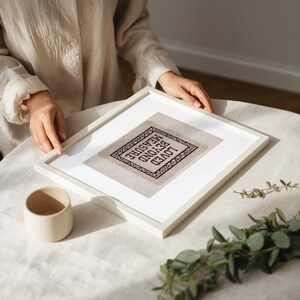 May include: A framed cross-stitch artwork with the words "LOVED BEYOND MEASURE" in a diamond shape. The artwork is in a white frame, and the cross-stitch is in black thread on a neutral fabric. A small, beige ceramic cup sits nearby.