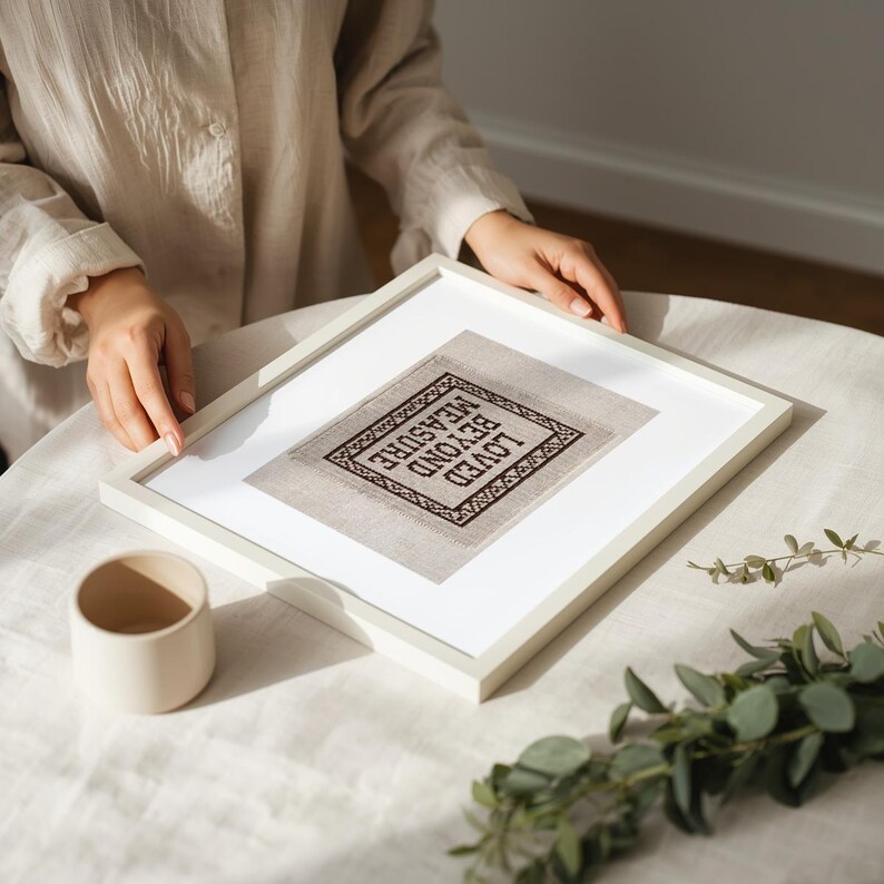 May include: A framed cross-stitch artwork with the words "LOVED BEYOND MEASURE" in a diamond shape. The artwork is in a white frame, and the cross-stitch is in black thread on a neutral fabric. A small, beige ceramic cup sits nearby.