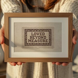 May include: A framed cross-stitch piece with the words "LOVED BEYOND MEASURE" in dark brown thread. The text is centered within a decorative border on a light beige fabric, set in a wooden frame with a white mat.