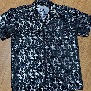 May include: Black button-up short-sleeve shirt with a flame pattern in white and blue. The shirt has a classic collar and a single chest pocket. The flame design covers the entire shirt, creating a bold and eye-catching look.