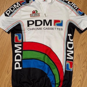 Puede incluir: Maillot de ciclismo blanco con detalles en negro y coloridos. La camiseta presenta el texto "PDM CHROME CASSETTES" y "ULTIMA". El diseño incluye un arco iris y el logo "PDM" en las mangas.