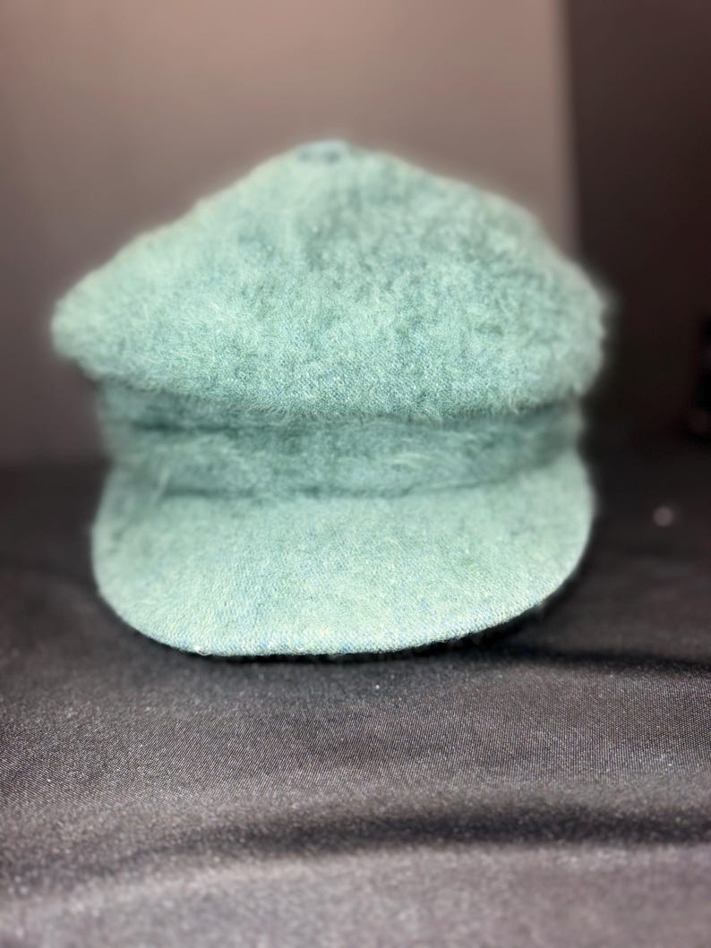 May include: A seafoam green newsboy cap with a rounded crown and a small, stiff brim. The hat has a textured, fuzzy appearance. The cap is made of a soft material and is suitable for casual wear.