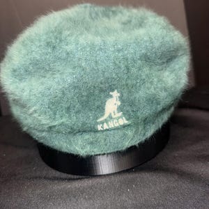 May include: A teal-colored beret with a soft, fuzzy texture. The beret features the white Kangol logo on the front. The hat is displayed on a black stand against a dark background.