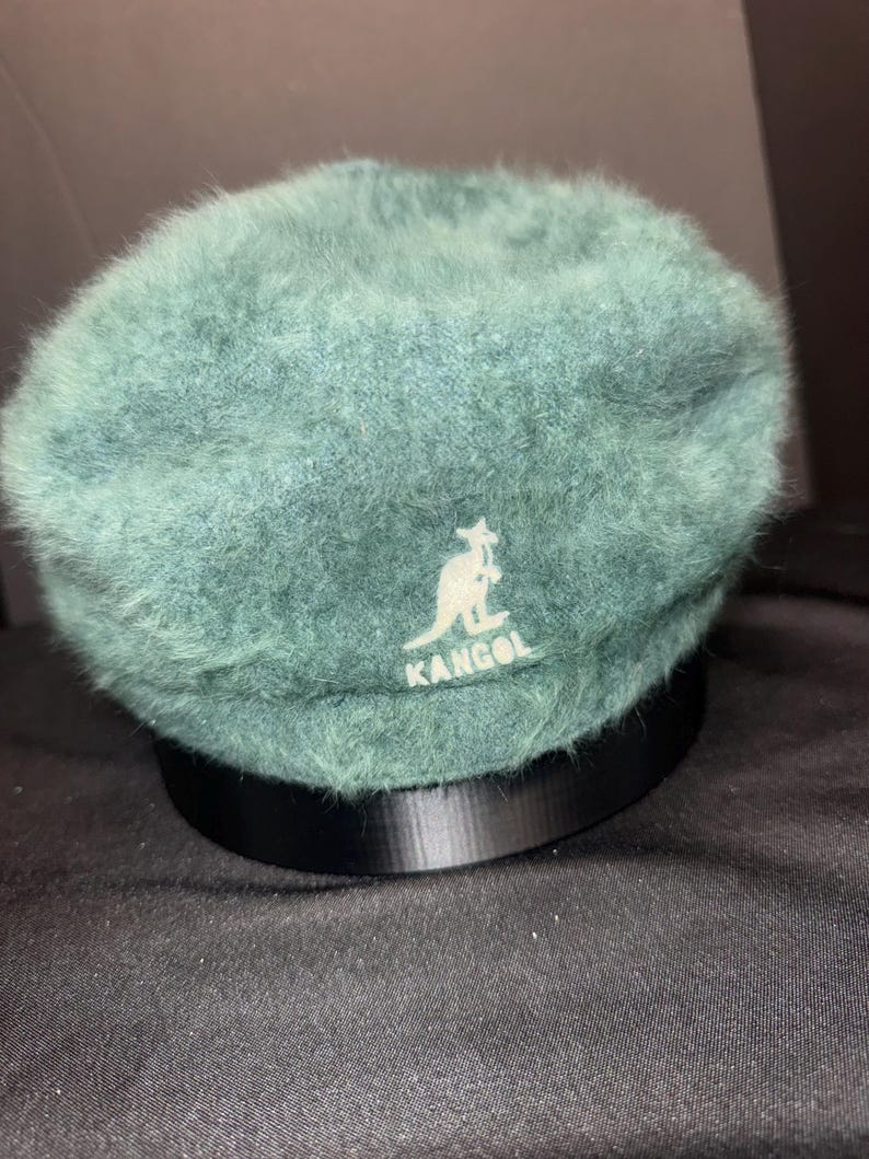 May include: A teal-colored beret with a soft, fuzzy texture. The beret features the white Kangol logo on the front. The hat is displayed on a black stand against a dark background.