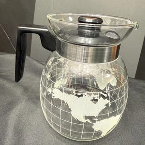 May include: A clear glass coffee carafe with a black handle and lid. The carafe is globe-shaped, with a world map etched onto the glass. The handle is black and the lid is dark brown.