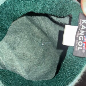 May include: A dark green Kangol hat with a soft, textured interior. The hat features a label with the Kangol logo and the Union Jack flag, indicating its origin in Great Britain. The hat's brim is a darker shade of green.