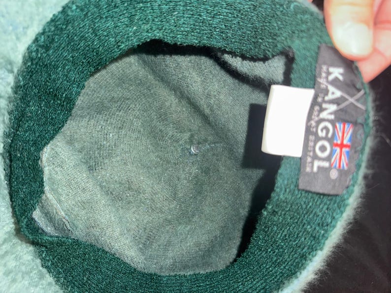 May include: A dark green Kangol hat with a soft, textured interior. The hat features a label with the Kangol logo and the Union Jack flag, indicating its origin in Great Britain. The hat's brim is a darker shade of green.