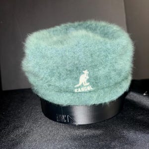 May include: A teal Kangol hat with a fuzzy texture. The hat features the Kangol logo in white and sits on a black stand. The hat has a rounded crown and a short brim.