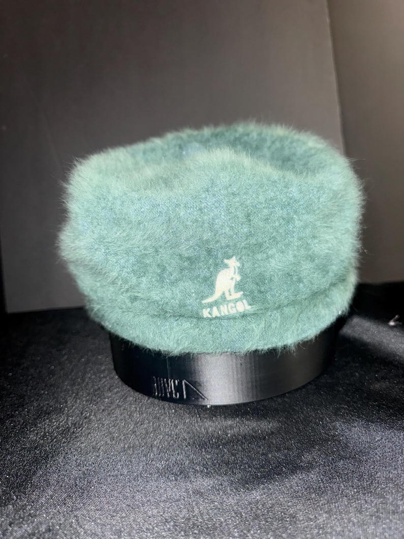 May include: A teal Kangol hat with a fuzzy texture. The hat features the Kangol logo in white and sits on a black stand. The hat has a rounded crown and a short brim.
