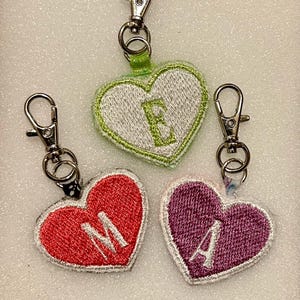 May include: Three heart-shaped keychains with embroidered letters. One is red with a white "M", another is purple with a white "A", and the third is green with a white "E". Each keychain has a silver-colored clasp.