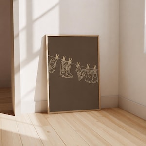 May include: A framed print featuring a brown background with a line drawing of cowboy-themed items hanging on a clothesline. The items include a cowboy hat, boots, a bandana, and denim shorts, all in a simple line art style.