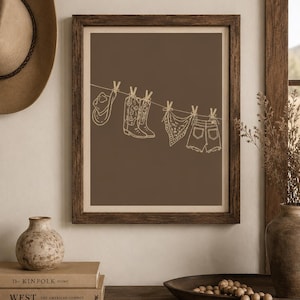 May include: Framed art print featuring a brown background with a line of cowboy-themed items. A cowboy hat, boots, a bandana, and denim shorts are depicted hanging on a clothesline. The artwork is in a minimalist style with a neutral color palette.