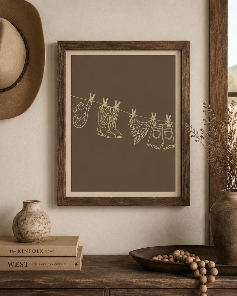 May include: Framed art print featuring a brown background with a line of cowboy-themed items. A cowboy hat, boots, a bandana, and denim shorts are depicted hanging on a clothesline. The artwork is in a minimalist style with a neutral color palette.