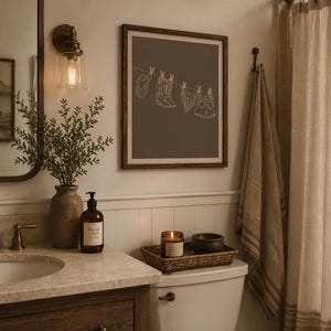 May include: A bathroom scene with a framed print featuring a line drawing of cowboy-themed items hanging on a clothesline. The room includes a vanity with a mirror, a lit candle, and a decorative towel.