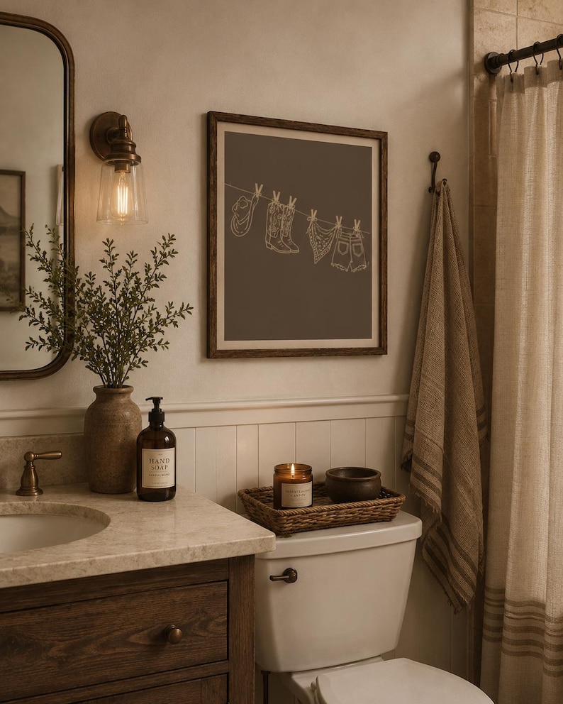 May include: A bathroom scene with a framed print featuring a line drawing of cowboy-themed items hanging on a clothesline. The room includes a vanity with a mirror, a lit candle, and a decorative towel.