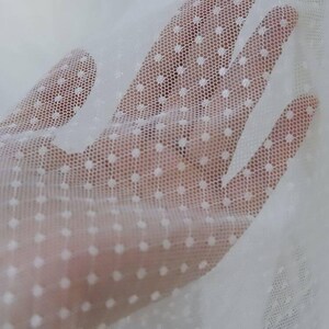 May include: Close-up of white sheer fabric with a polka dot pattern. The fabric is held up, revealing the texture and transparency. The dots are evenly spaced and add a delicate touch to the material.