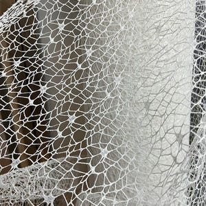 May include: Close-up of white lace fabric with a delicate, open net pattern. The fabric is tightly woven, creating a web-like design. The texture is visible, with a slight sheen. The fabric is likely used for clothing or decorative purposes.