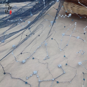 Puede incluir: Tela de tul azul oscuro con constelaciones bordadas, pequeños detalles de flores azules y lentejuelas dispersas. La tela es semitransparente, con una apariencia delicada y aireada. El bordado crea un patrón celestial.