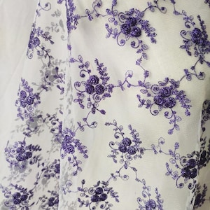 Könnte beinhalten: Nahaufnahme eines weißen, transparenten Stoffes mit einem sich wiederholenden Blumenmuster in Lilatönen. Das Design zeigt detaillierte gestickte Blumen und Blätter, die ein zartes und strukturiertes Aussehen erzeugen. Ideal für Bastelarbeiten oder Kleidung.