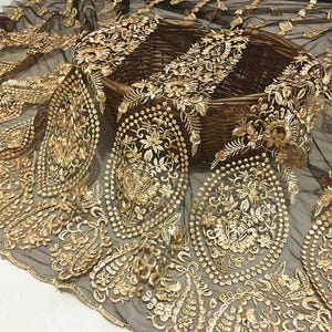 May include: Black lace fabric with intricate gold floral embroidery. The detailed design features ornate patterns and a delicate, sheer texture. The fabric is draped over a woven basket, showcasing the luxurious gold threadwork.