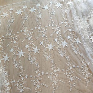 Könnte beinhalten: Weißer Stoff mit Sternenmuster. Der Stoff weist ein filigranes Design aus unterschiedlich großen Sternen auf, das ein himmlisches Thema erzeugt. Die Sterne sind aufwendig gestickt und verleihen dem Material Textur und Dimension. Ideal für Bastelarbeiten oder Dekorationszwecke.