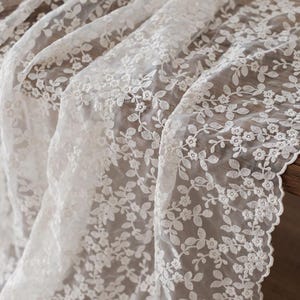 Puede incluir: Primer plano de una tela de encaje blanco con un diseño floral. El delicado encaje presenta pequeñas flores y hojas, creando un diseño texturizado y elegante. La tela está drapeada, mostrando su calidad fluida y sus intrincados detalles.