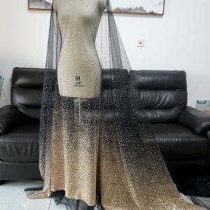 Pode incluir: Tecido de tule preto com um gradiente de glitter dourado, passando de preto no topo para dourado na parte inferior. O tecido é drapeado sobre um manequim, mostrando sua textura e brilho. Ideal para roupas de noiva ou formais.