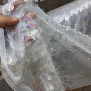 Könnte beinhalten: Nahaufnahme eines weißen Stoffes mit schillernden Pailletten. Die runden Pailletten reflektieren ein Regenbogenspektrum an Farben. Der Stoff wird gehalten, wodurch seine Textur und sein Fall sichtbar werden.