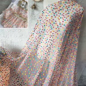 Puede incluir: Tela blanca transparente con un estampado de lunares coloridos en tonos rojos, amarillos, verdes, azules y morados. La tela está drapeada sobre un maniquí, mostrando su textura fluida. Una imagen más pequeña muestra un vestido hecho con la misma tela.