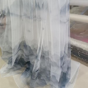 以下が含まれることがあります： グレーとブルーの色合いで水彩画風の山脈模様が描かれた、透け感のある白い生地。生地はドレープされており、デザインのレイヤー効果を示しています。素材は軽く、流れるように見えます。