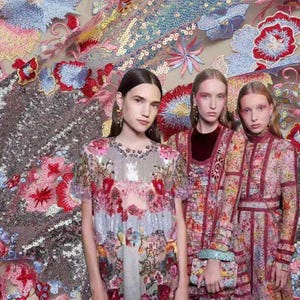 Pode incluir: Três mulheres em vestidos e tops bordados com flores. Os vestidos apresentam intrincados padrões florais em vermelho, rosa, azul e amarelo. O fundo é um tecido de lantejoulas com bordados florais.