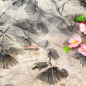 以下が含まれることがあります： 黒い刺繍の花柄が施されたシアー生地のクローズアップ。生地は淡いベージュ色で、刺繍は詳細な枝のような模様が特徴です。ピンクの花と緑の葉が右上隅にあります。