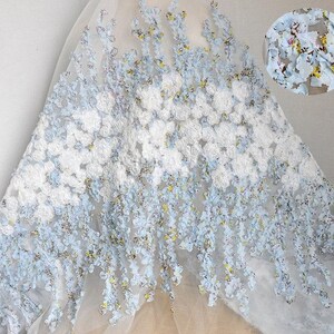 Puede incluir: Delicada tela de encaje floral blanco y azul claro. El encaje presenta racimos de flores blancas y acentos florales azules en cascada con detalles amarillos. Ideal para vestidos de novia, vestidos o proyectos decorativos.