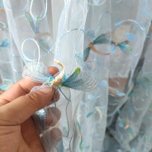 Puede incluir: Cortina transparente azul claro con diseño de libélulas bordadas. La libélula tiene detalles en azul, dorado y verde. La cortina está hecha de una tela delicada y semitransparente, con múltiples libélulas bordadas en la superficie.