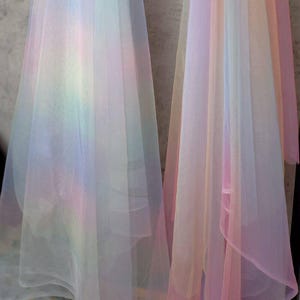 Pode incluir: Dois painéis de tecido transparente em um gradiente de arco-íris suave. As cores misturam-se de rosa pastel e amarelo a azul e verde. O tecido parece leve e fluido, adequado para fins decorativos.
