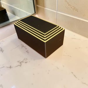 May include: A rectangular black box with a stepped, tiered top design. The edges of each tier are highlighted with a thin, light-colored trim, creating a layered effect. The object has a solid, matte black finish on its sides and top surfaces.