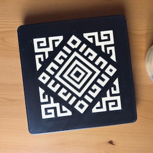 Puede incluir: Una caja decorativa cuadrada negra con un patrón geométrico blanco. El diseño presenta una forma de diamante central rodeada por una serie de cuadrados y rectángulos entrelazados. La caja está sobre una superficie de madera.