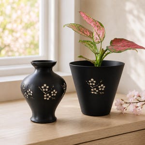 May include: A matte black vase with a narrow neck and a wide body featuring carved white cherry blossom motifs, paired with a matching matte black tapered plant pot containing a plant with pink and green leaves and a white cherry blossom design.