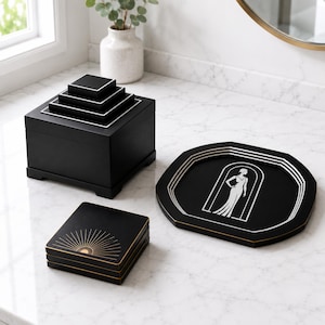 Pode incluir: Uma caixa decorativa preta com tampa em camadas, uma bandeja preta com um design art déco prateado e branco e um conjunto de porta-copos pretos com um padrão de sol dourado. Os itens são exibidos em uma superfície de mármore branco.