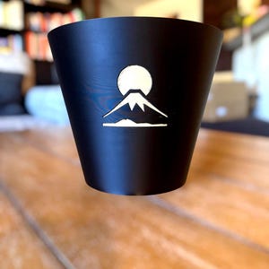 May include: Black cup with a white mountain and sun design. The cup has a tapered shape and a smooth surface. The design is a simple, minimalist illustration. The cup is sitting on a wooden surface.
